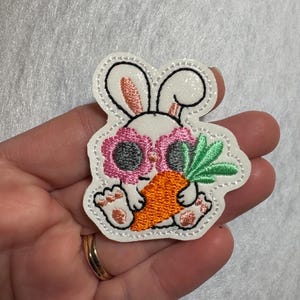 May include: Embroidered patch featuring a white bunny wearing pink sunglasses and holding an orange carrot with green leaves. The patch has a white border with black stitching and is designed in a cartoon style.