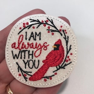 May include: White glitter circle with a red cardinal bird and a black embroidered wreath with red berries. The text "I AM always WITH YOU" is embroidered in black.