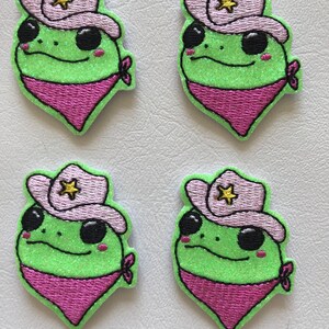 May include: Four embroidered patches featuring a cartoon frog wearing a pink cowboy hat with a gold star and a pink bandana. The frog is green with black eyes and a glittery texture.