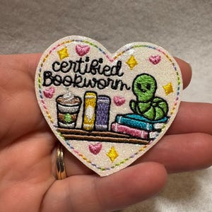 May include: Heart-shaped patch with the words "certified Bookworm" embroidered in black. The patch features a coffee cup, books, and a green bookworm. Pink hearts and yellow stars decorate the border.
