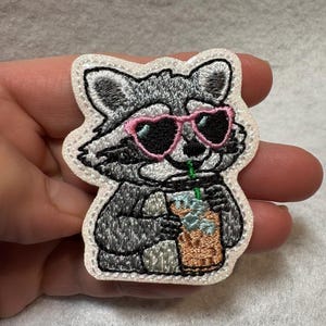 May include: Embroidered patch featuring a raccoon wearing pink heart-shaped sunglasses, holding a beverage with a green straw. The raccoon is gray and white, with a white border. The patch is designed for application on clothing or accessories.