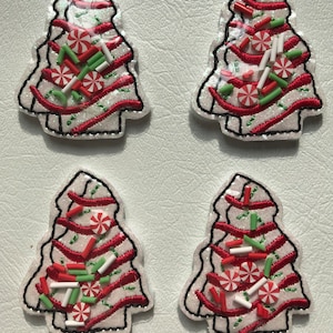 May include: Four embroidered patches shaped like Christmas trees, featuring white glittery fabric with red stitched swirls. Each patch is decorated with small, flat, circular peppermint candy and cylindrical sprinkle embellishments in red, white, and green.