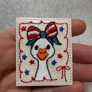 May include: Embroidered patch featuring a white goose with a red, white, and blue bow. The patch has a glittery background with red and blue star accents and a red stitched border. The goose has an orange beak.