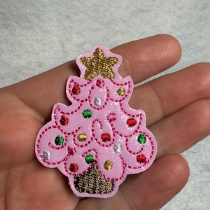 May include: A pink glittery Christmas tree patch featuring embroidered details. The tree is topped with a gold star and decorated with small, colorful embroidered circles in red, green, silver, and gold. The base of the tree is dark brown.