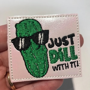 May include: A white, square, embroidered patch with a green pickle wearing sunglasses and the text "JUST DILL WITH IT!"