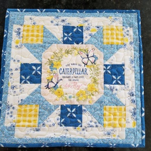 Peut inclure: Une suspension murale matelassée bleue et jaune avec un carré de tissu blanc au centre. Le carré blanc présente un motif floral et le texte "Just when the caterpillar thought it was over, she began to fly."