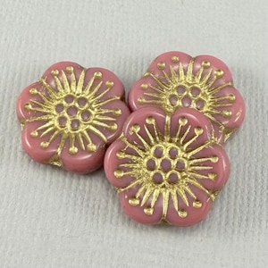 May include: Three pink flower-shaped beads with gold-colored accents. The beads have a raised floral design with a central cluster of dots and radiating lines. The beads are arranged on a white surface.