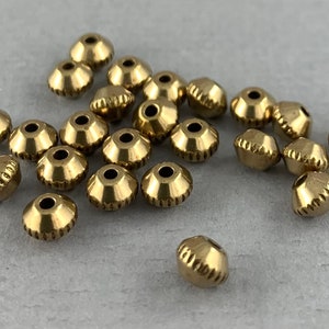 May include: A group of 20 gold-colored metal beads with a decorative, ridged design. The beads are scattered on a gray surface.