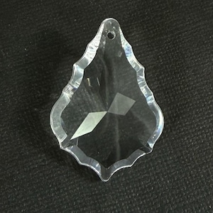 May include: Clear, faceted crystal teardrop pendant with a scalloped edge and a small hole at the top for hanging. The crystal reflects light, creating a sparkling effect. Ideal for chandeliers or decorative projects.