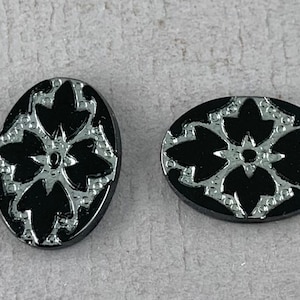 May include: Two black oval buttons with a silver floral design. The buttons have a raised, textured surface.