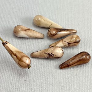 May include: Six teardrop-shaped glass beads with a brown and white marbled pattern. The beads are approximately 2.5 cm long.