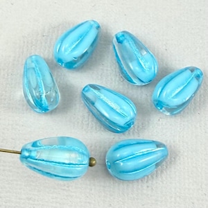 Könnte beinhalten: Eine Sammlung tropfenförmiger Perlen in Hellblau und transparentem Glas. Jede Perle hat ein geripptes Design und ein Loch zum Auffädeln. Die Perlen liegen auf einer weißen Oberfläche, wobei eine Perle an einer goldfarbenen Nadel befestigt ist.