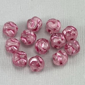 May include: A collection of pink, swirled beads. The beads have a glossy finish and a marbled appearance, with shades of pink and white. The beads are round and have a slightly irregular shape. The beads are scattered on a white surface.