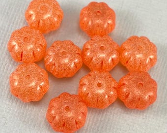 Puede incluir: Una colección de diez cuentas naranjas con forma de flor. Cada cuenta tiene un agujero central y un diseño texturizado en forma de pétalo. Las cuentas están dispuestas sobre una superficie blanca, mostrando su color vibrante y forma única.