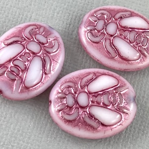 May include: Three oval-shaped glass beads with a pink iridescent finish. Each bead features a raised white design of a stylized insect.