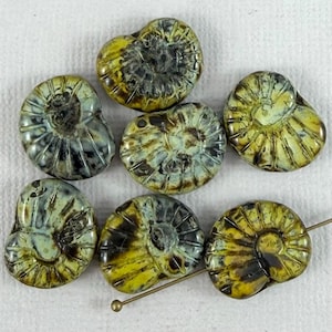Op de afbeelding: Een verzameling van zeven fossielvormige kralen. De kralen zijn een mix van geel, groen en zwart, met een spiraalvormig ontwerp. Een goudkleurige pin is door een van de kralen gestoken. De kralen liggen op een witte ondergrond.