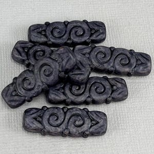 May include: A collection of six dark purple rectangular beads. Each bead features a repeating pattern of swirling designs. The beads are arranged on a white surface. The beads are likely for jewelry making or crafting.