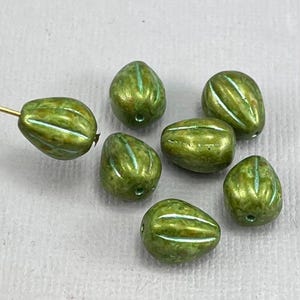 Könnte beinhalten: Sieben grüne und weiße, gestreifte Glasperlen mit Perlmutt-Finish. Die Perlen sind oval geformt und haben einen dezenten Glanz.