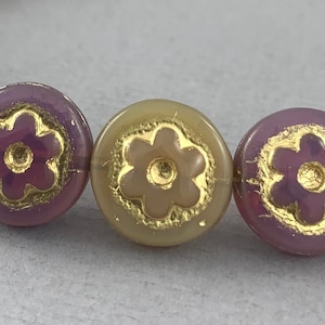 May include: Close-up of three round glass beads with a floral design. The beads are a light purple and beige color with gold accents.