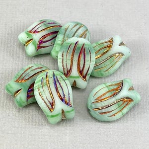 May include: A collection of light green glass beads shaped like tulip buds. Each bead features gold and iridescent accents, with vertical lines adding detail. The beads are clustered together, showcasing their unique design and color variations.
