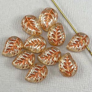 May include: Ten translucent glass beads in the shape of leaves with a copper-coloured leaf design. The beads are scattered on a white surface.