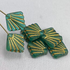 May include: A group of teal green glass beads with a gold fan design. The beads are square shaped and have a frosted finish.