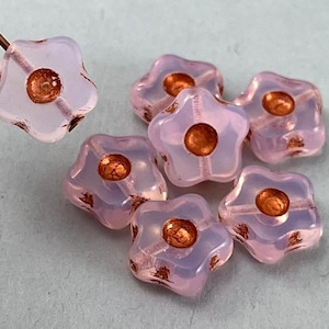 May include: A group of six pale pink glass flower-shaped beads with copper accents. The beads have a star-shaped outline and a small copper circle in the center.