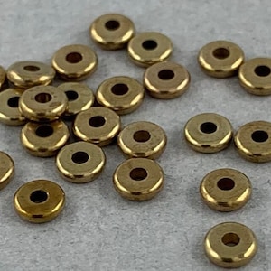 May include: A collection of small, round, gold-colored metal beads with a hole in the center. The beads are scattered on a light gray surface. These beads are likely used for jewelry making or other craft projects.