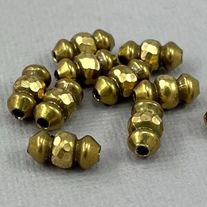May include: A collection of small, gold-coloured metal beads. Each bead has a faceted, textured surface and a cylindrical shape with a hole at each end. The beads are scattered on a light grey surface.