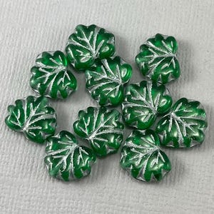 May include: A collection of green glass leaf-shaped beads with a white raised design. The beads are approximately 1 inch in size.