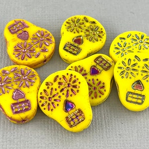 May include: Seven yellow glass skull beads with pink and gold accents. The beads are decorated with floral patterns and have a heart shape in the centre of the skull.