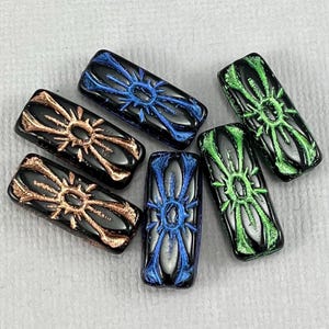 May include: Six rectangular glass beads with a black base and a raised, stylized sunburst design in copper, blue, and green. The beads are arranged on a light gray surface, showcasing their glossy finish and intricate details.
