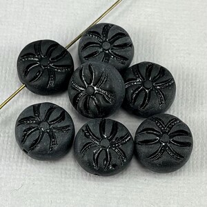 May include: Seven matte black glass beads with a floral design. Each bead is round with a slightly domed surface. The floral pattern is etched into the surface, creating a subtle contrast. A thin, gold-colored wire runs through some of the beads.