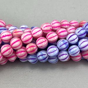 以下が含まれることがあります： ピンクとパープルのストライプが入った丸い白いビーズのストランド。ビーズはグラデーションパターンで配置されており、ピンクのビーズが片側に、パープルのビーズが反対側にあります。