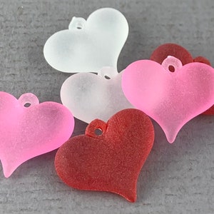 May include: A collection of heart-shaped charms in various colors: white, light pink, and red. Each charm has a small loop at the top for attaching to jewelry or other crafts. The charms have a frosted, textured appearance.