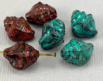 May include: Six seashell-shaped beads in shades of red and turquoise with black accents. The beads are arranged on a white surface, with one bead attached to a gold-colored pin. These beads are suitable for jewelry making.