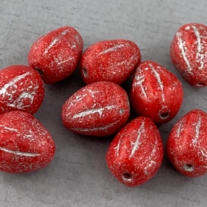 Könnte beinhalten: Eine Sammlung roter, tropfenförmiger Perlen mit silberfarbenen, strukturierten Streifen. Jede Perle hat ein kleines Loch zum Auffädeln. Die Perlen haben ein leichtes Vintage-Aussehen, geeignet für Schmuckherstellung oder Bastelarbeiten.