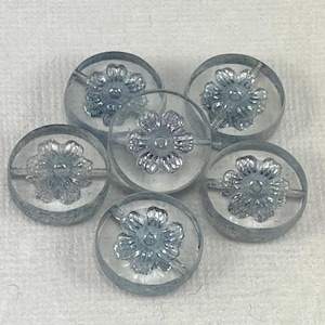 May include: Six clear, round buttons with a floral design in the center. The buttons have a light blue hue and are arranged in a circular pattern. Each button has two holes for sewing.