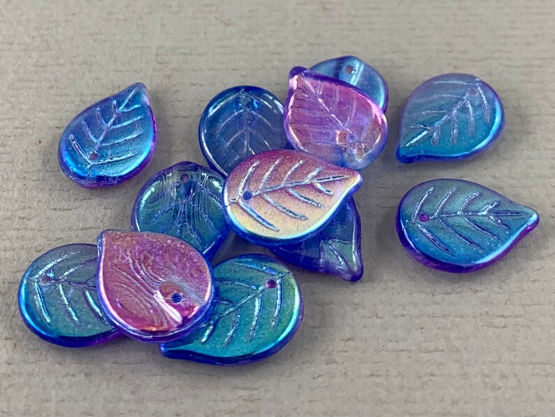 May include: A collection of iridescent leaf-shaped beads. The beads have a blue and purple color shift, with visible leaf vein details. Each bead has a small hole for stringing. These beads are suitable for jewelry making and craft projects.