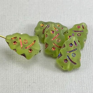 May include: A collection of light green, tree-shaped glass beads. Each bead features iridescent accents in pink, purple, and gold, with a frosted finish. The beads are designed with a textured surface and decorative details.