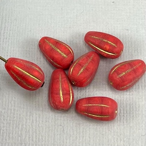 Könnte beinhalten: Rote, tropfenförmige Perlen mit Goldstreifen. Die Perlen sind auf einer weißen Oberfläche angeordnet. Die Perlen haben eine matte Oberfläche und sind wahrscheinlich aus Glas oder Keramik. Die Perlen sind für die Schmuckherstellung.