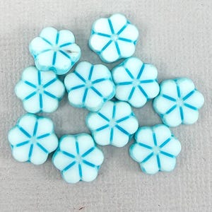 Peut inclure: Dix perles en forme de fleur. Chaque perle est blanche avec un contour bleu clair et des lignes bleues partant du centre. Les perles sont dispersées sur une surface gris clair.