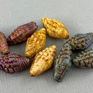 Op de afbeelding: Assortiment van tien keramische kralen in verschillende kleuren en patronen. De kralen zijn gevormd als kleine schelpen met een getextureerd oppervlak. De kleuren omvatten paars, bruin, crème en blauw.