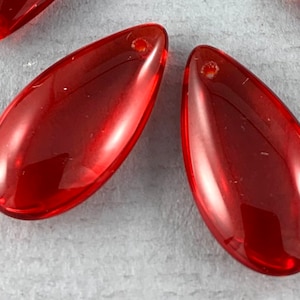 May include: Close-up of several transparent, teardrop-shaped beads in a vibrant red hue. Each bead has a small hole at the top for stringing. The beads are smooth and glossy, reflecting light.