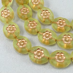 Peut inclure: Gros plan sur un rang de perles ovales vert clair, chacune ornée d'un motif floral doré au centre. Les perles sont enfilées sur une fine chaîne dorée, créant un bijou.