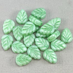 May include: A collection of light green glass beads shaped like leaves. The beads have a raised vein pattern and a glossy finish.
