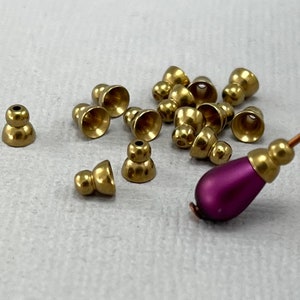 May include: A group of 15 gold-coloured metal bead caps, some with a rounded top and some with a flat top. The bead caps are scattered on a white surface. A purple bead is in the bottom right corner of the image.