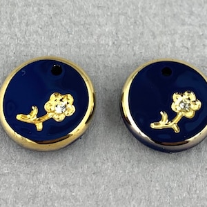 Könnte beinhalten: Zwei runde Anhänger mit goldfarbenem Rand, blauem Emaille-Hintergrund und einer goldenen Blume mit einem klaren Strassstein in der Mitte.