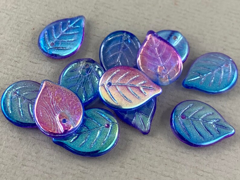 May include: A collection of iridescent glass leaf-shaped beads. The beads are primarily blue with hints of purple and gold, and feature a detailed leaf vein design. Each bead has a small hole for stringing.