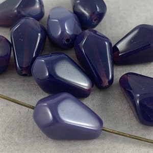 May include: A collection of dark blue, teardrop-shaped beads with a glossy finish. Each bead has a small hole for stringing and a subtle, darker stripe. A brass-colored pin is inserted through one of the beads.